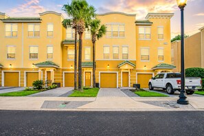 A yellow multi-story building with multiple garage doors, a white pickup truck parked in front, and a well-maintained lawn. - Spacious 3BR Townhouse in Vista Cay – Sleeps 8 Near Disney (Orlando)