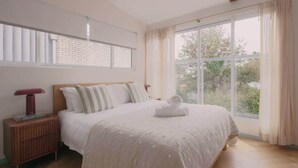 A bedroom with a large window, a bed with a white bedspread, a nightstand with a lamp, and a brick wall. - BONDI SALT & SURF 3 bedroom apartment with Parking (Bondi Beach)