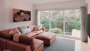 A modern living room with a sectional sofa, a large window, and a painting on the wall. - BONDI SALT & SURF 3 bedroom apartment with Parking (Bondi Beach)