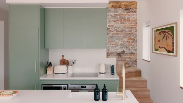 A modern kitchen with green cabinets, a brick accent wall, and a staircase leading to another level. - BONDI SALT & SURF 3 bedroom apartment with Parking (Bondi Beach)