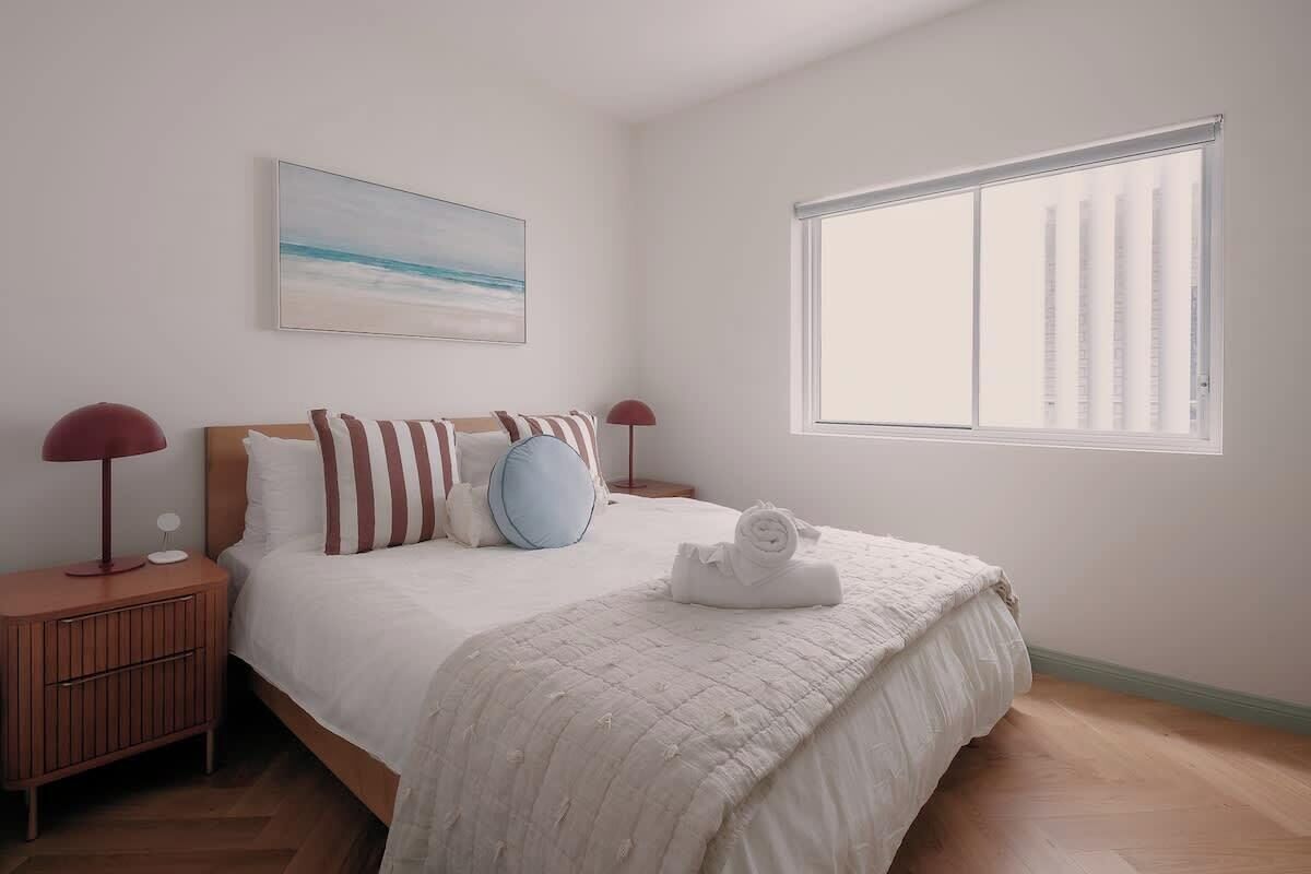 Ein Schlafzimmer mit einem Bett, einem Nachttisch, einer Lampe, einem Fenster und einem Bild an der Wand.