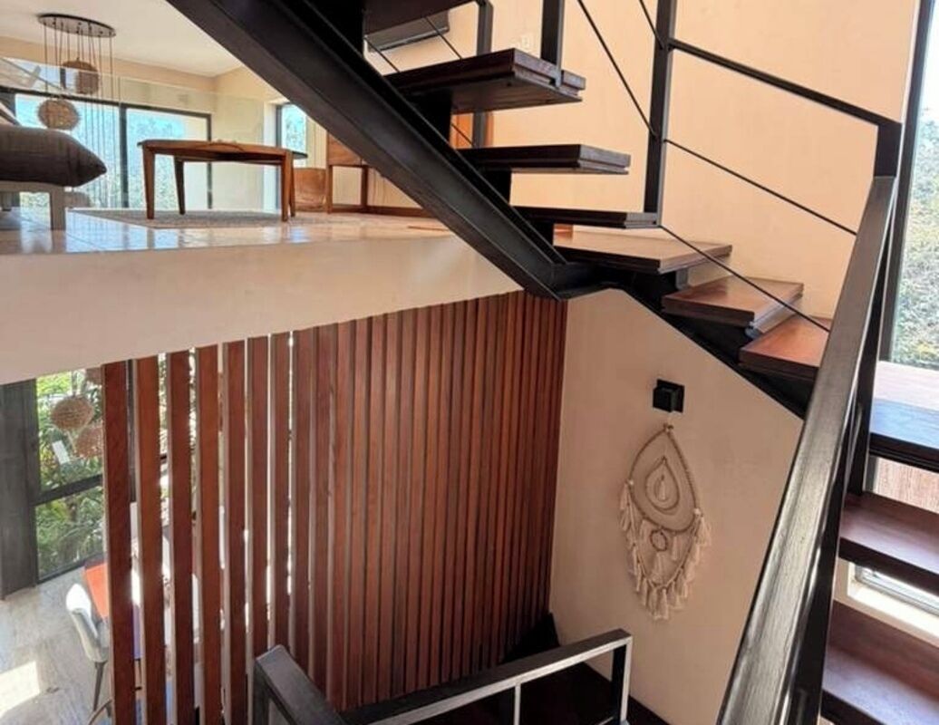A modern staircase with a wooden finish and metal railings, leading to a high-ceilinged area with a view of the outdoors.