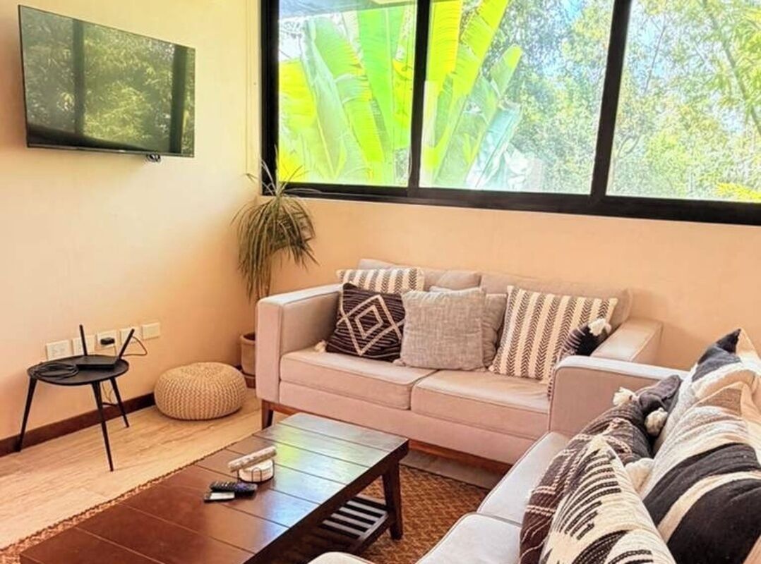 A living room with a sofa, a coffee table, a TV mounted on the wall, and a large window with a view of greenery.
