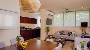 Een moderne woonkamer met een bank, salontafel en eettafel. Er is een keuken met donkerhouten kasten en een groot raam.