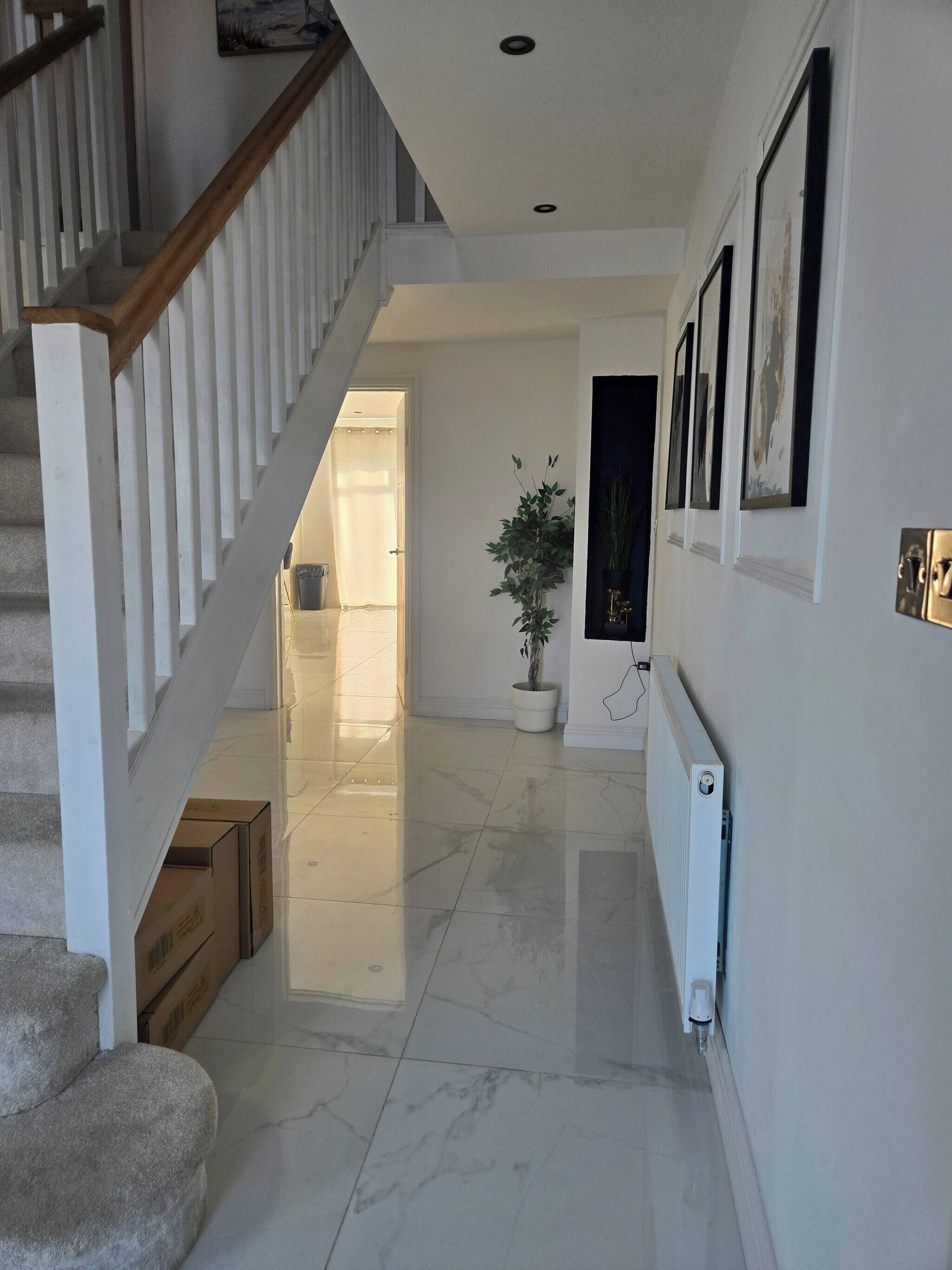 Un pasillo con piso de mármol, una escalera con barandillas blancas, una planta en maceta y cuadros en la pared.