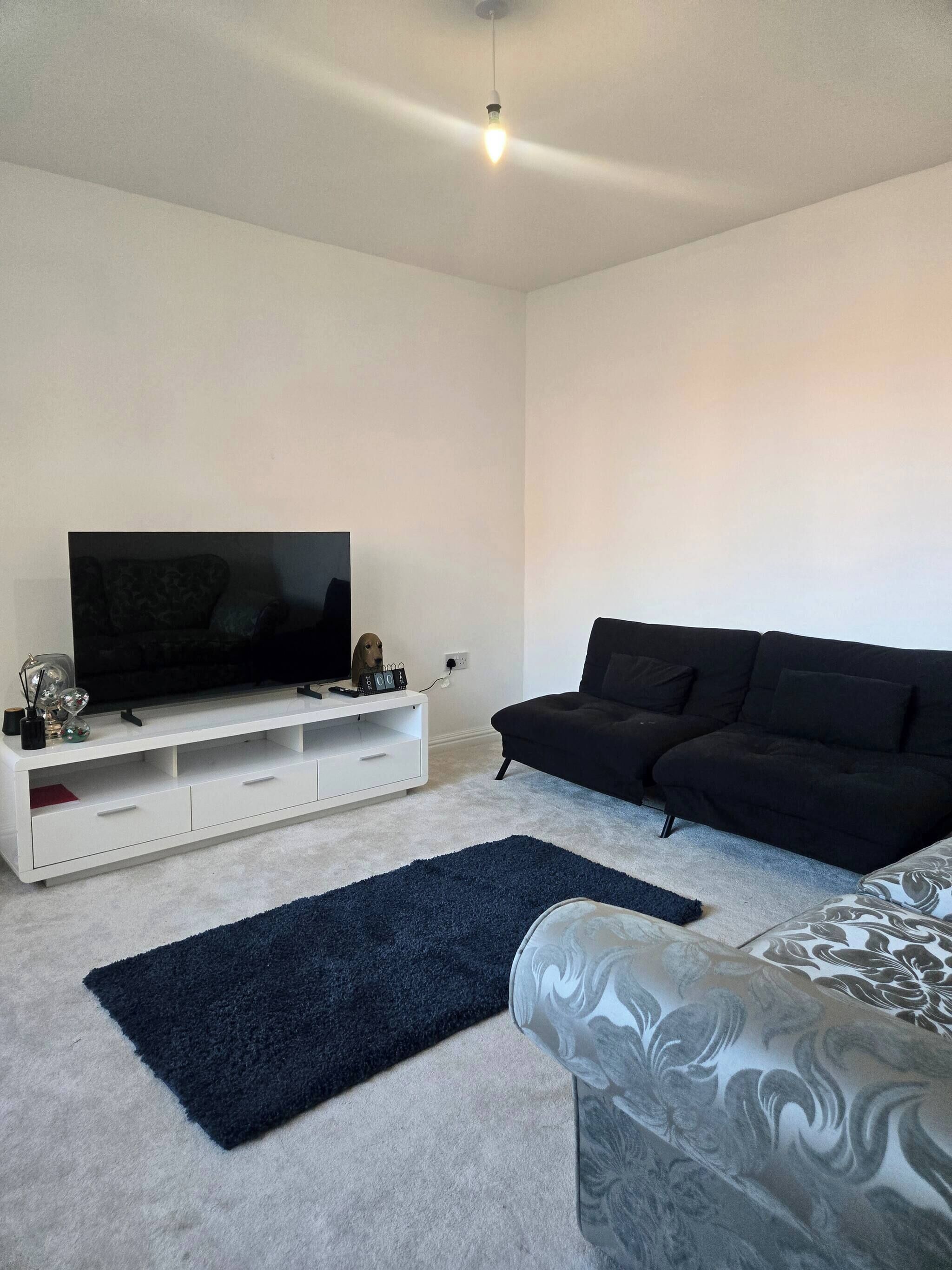 A modern living room with a flat-screen TV on a white entertainment unit, a black sofa, a patterned armchair, and a dark blue rug.