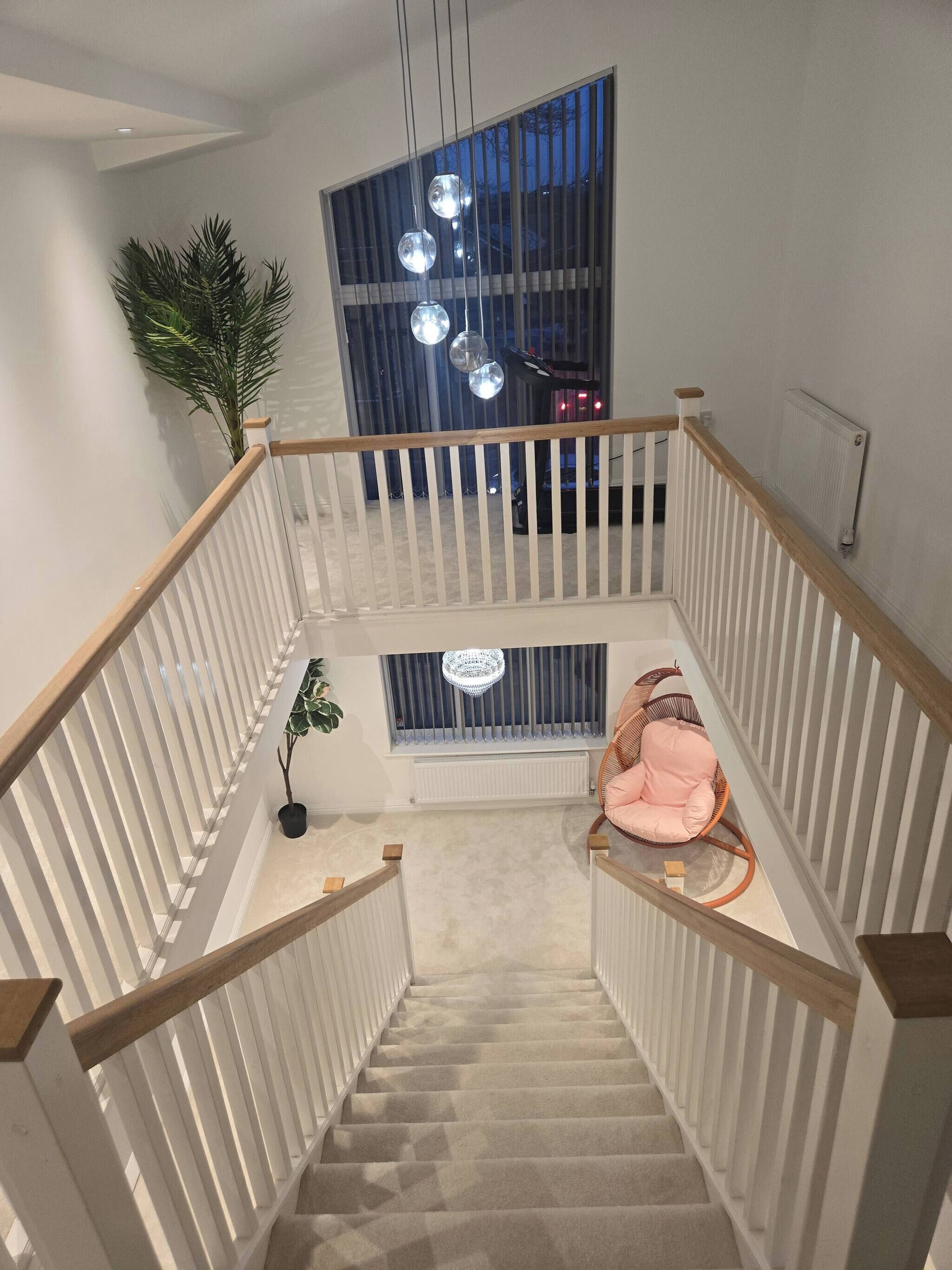 A staircase with white railings and a wooden handrail, leading to a landing with a large window, a chandelier, and a pink chair.