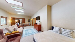 Ein Schlafzimmer mit einem Bett, einem Sofa, einer Kommode und einem Fernseher. Im Hintergrund ist ein Essbereich mit Tisch und Stühlen zu sehen.