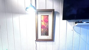 Ein Zimmer mit einem Holztisch und zwei roten Stühlen, einem gerahmten Gemälde an der Wand und einem an der Wand befestigten Fernseher.
