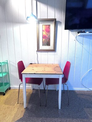 A room with a wooden table and two red chairs, a framed painting on the wall, and a television mounted on the wall. - Point Charming Studio (Richmond)