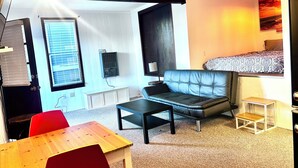 Ein modernes Wohnzimmer mit einer Ledersofa, einem Holztisch, einem roten Sessel, einem kleinen Holzschemel und einem Bett mit gemusterten Bettbezügen.