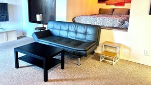 Ein modernes Wohnzimmer mit einer Ledersofa, einem schwarzen Couchtisch, einem kleinen Holznachttisch und einem Bett mit gemusterten Bettbezügen.