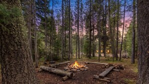 A forest campsite with a central fire pit surrounded by logs, dense trees, and a distant building. - Adventure Creek Lodge | Hot Tub, Zipline & Kayaks (Island Park)