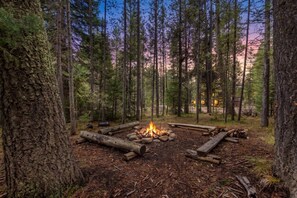 A forest campsite with a central fire pit surrounded by logs, dense trees, and a distant building. - Adventure Creek Lodge | Hot Tub, Zipline & Kayaks (Island Park)
