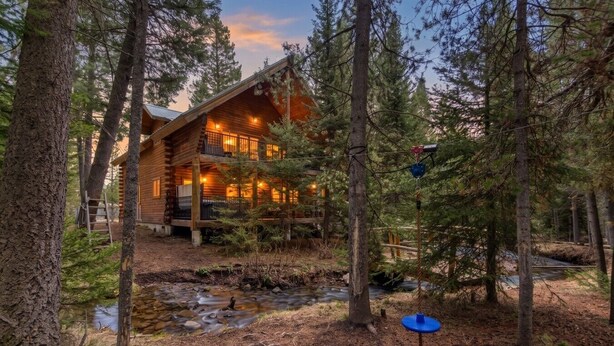 A log cabin with a porch, surrounded by trees and a stream. - Adventure Creek Lodge | Hot Tub, Zipline & Kayaks (Island Park)