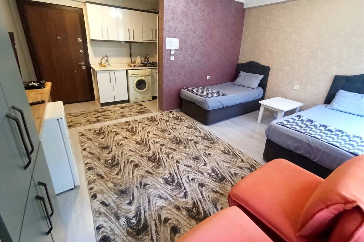 A compact room with a bed, a small table, a sofa, a washing machine, and a kitchenette.