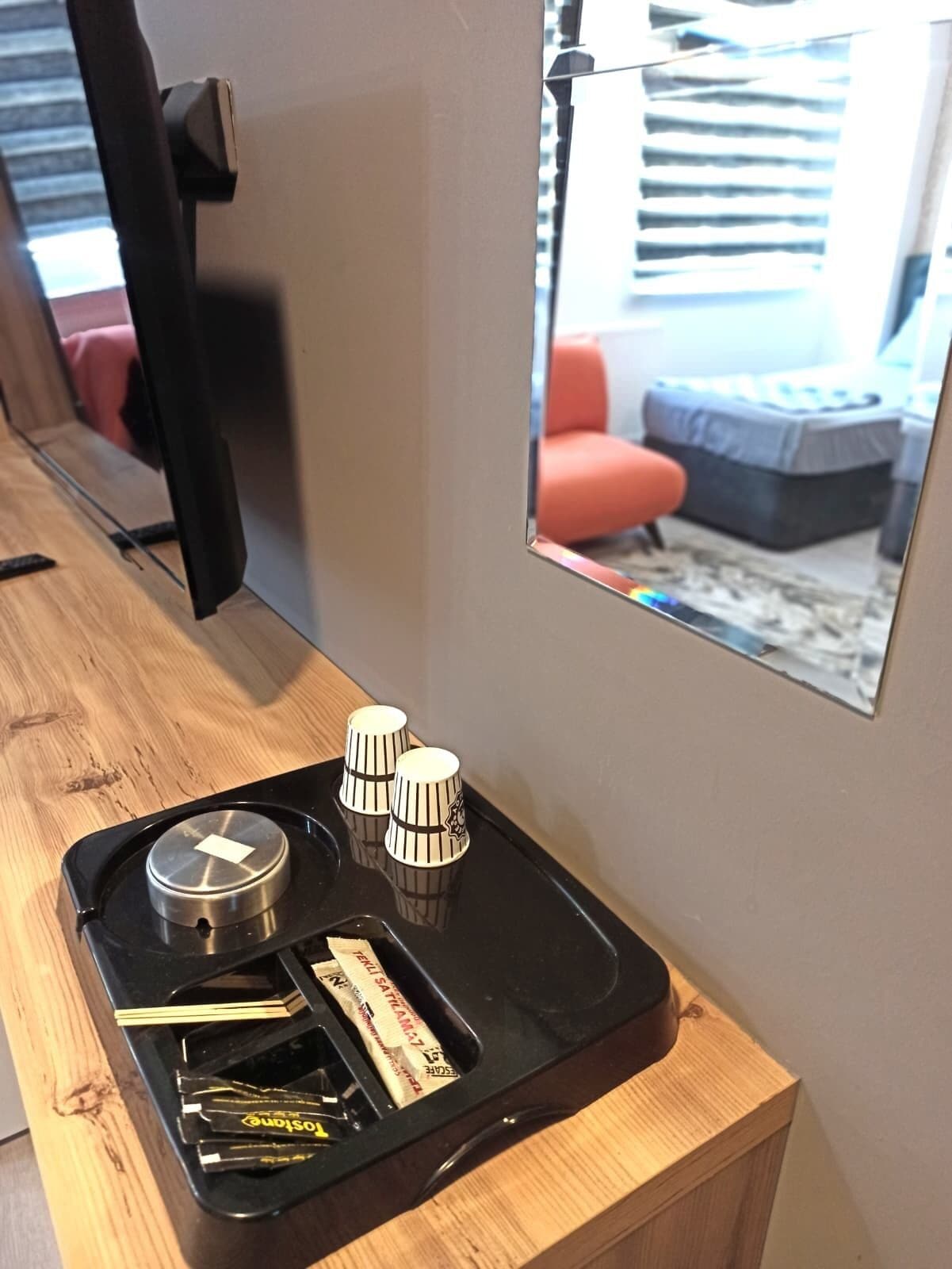 A hotel room with a wooden desk, a black tray with a bottle, a pack of tissues, and a small mirror reflecting a red chair and a gray sofa.