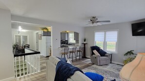 A spacious living room with a grey sofa, a coffee table, and a TV. A kitchen area with a microwave and a dining area with a table and chairs is visible in the background.