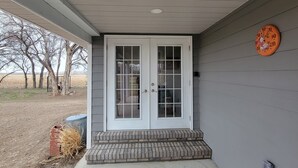 A house with a front door, a small porch, a clock on the wall, and a view of trees and a field.