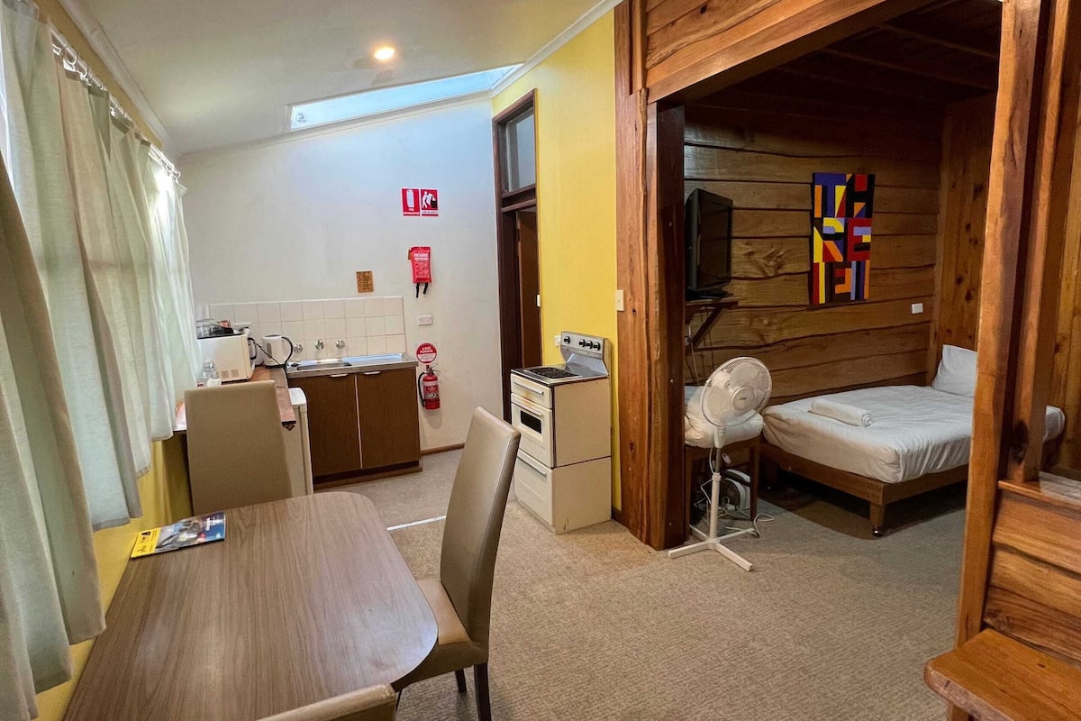 A compact hotel room with a dining table, a single bed, a small kitchenette, and a wooden wall decoration.