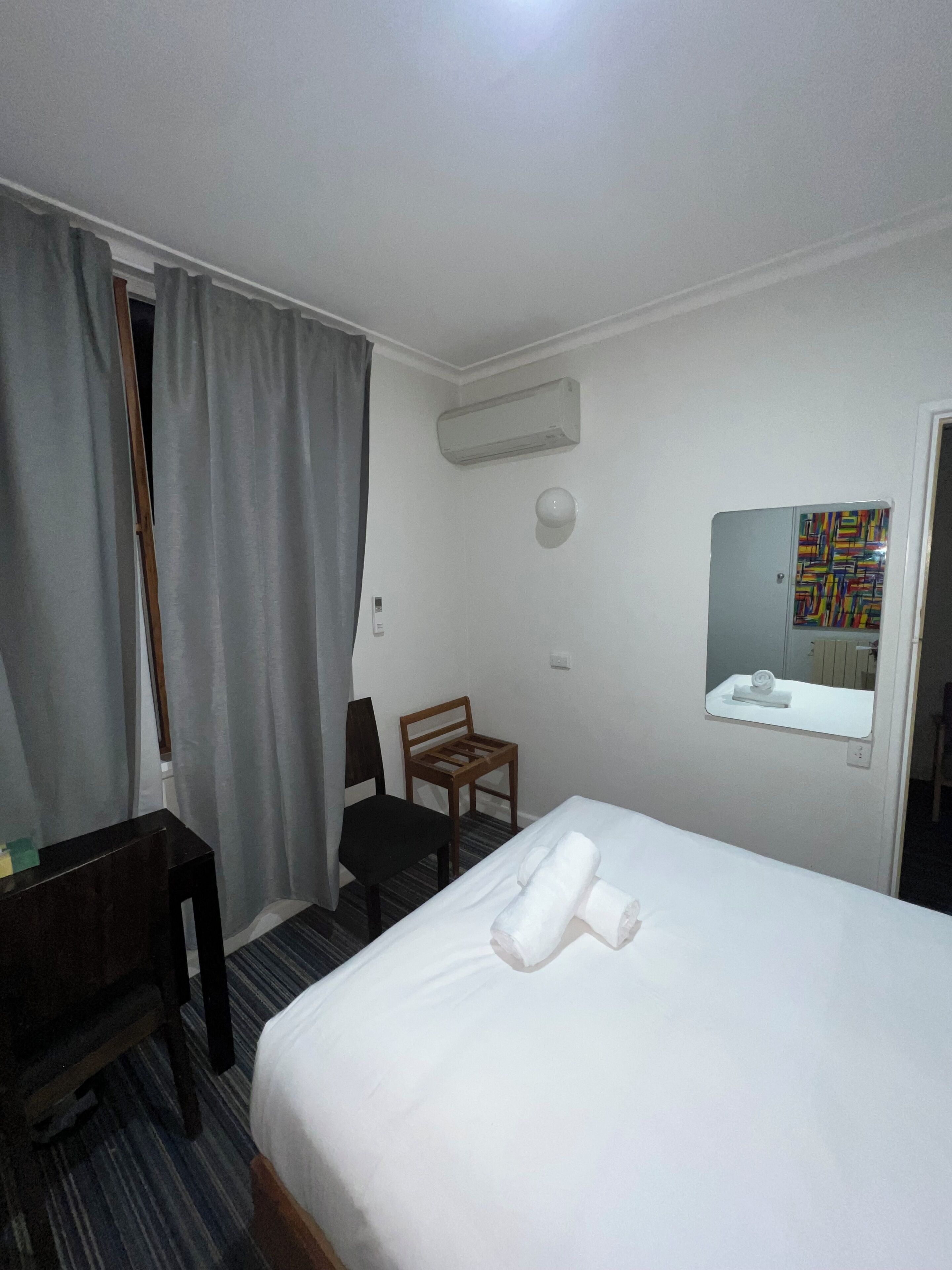 A hotel room with a bed, a desk, a chair, a mirror, and an air conditioning unit.