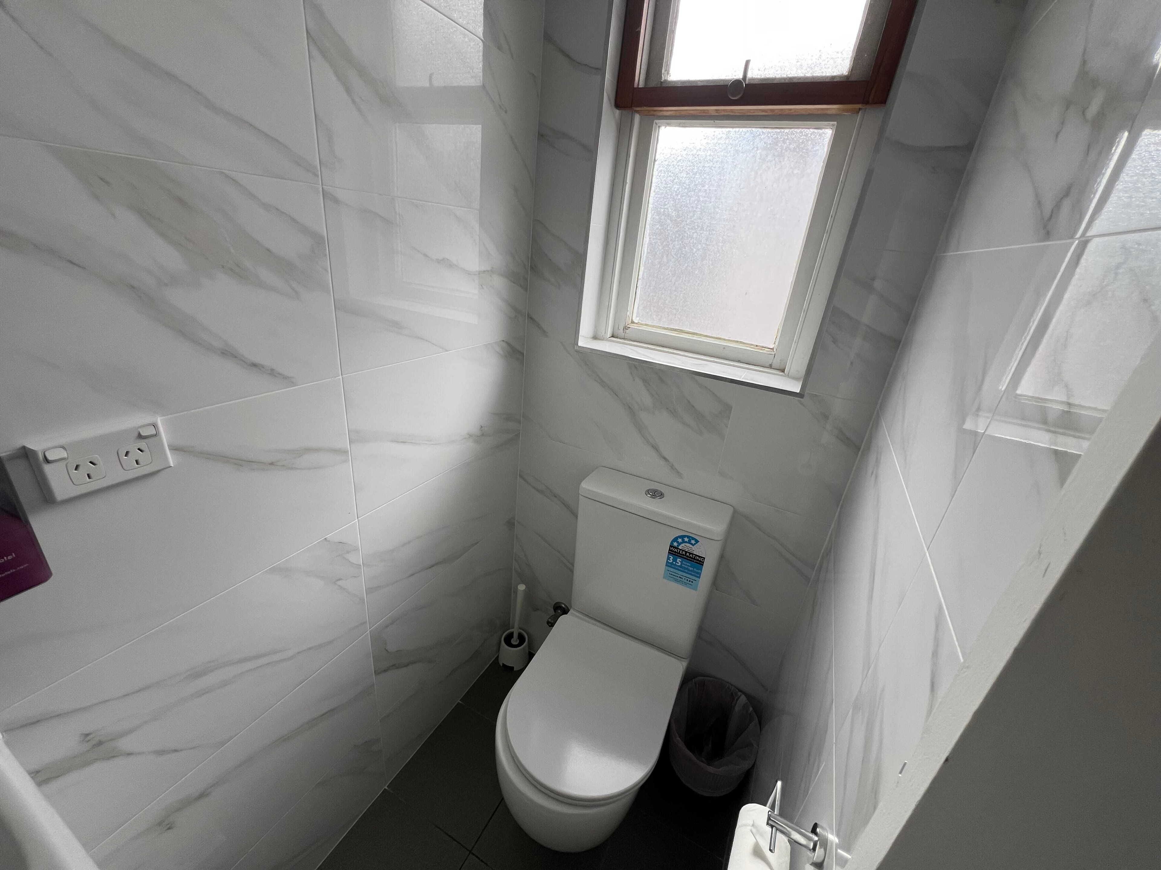 A small bathroom with a toilet, a window, and a trash bin.