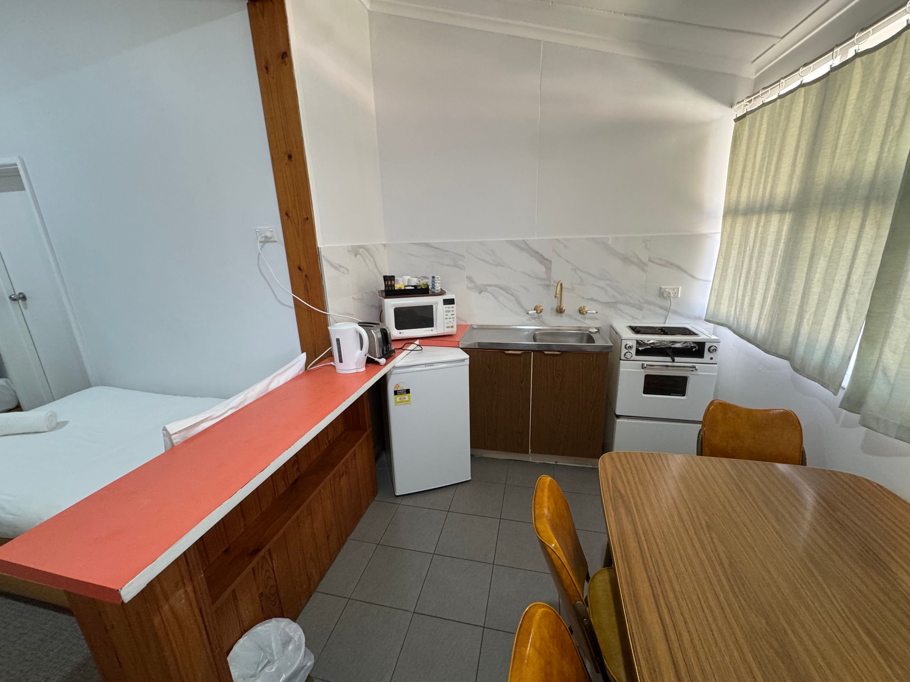 Quarto de hotel compacto com cozinha compacta, uma cama, mesa pequena e janela com cortinas.