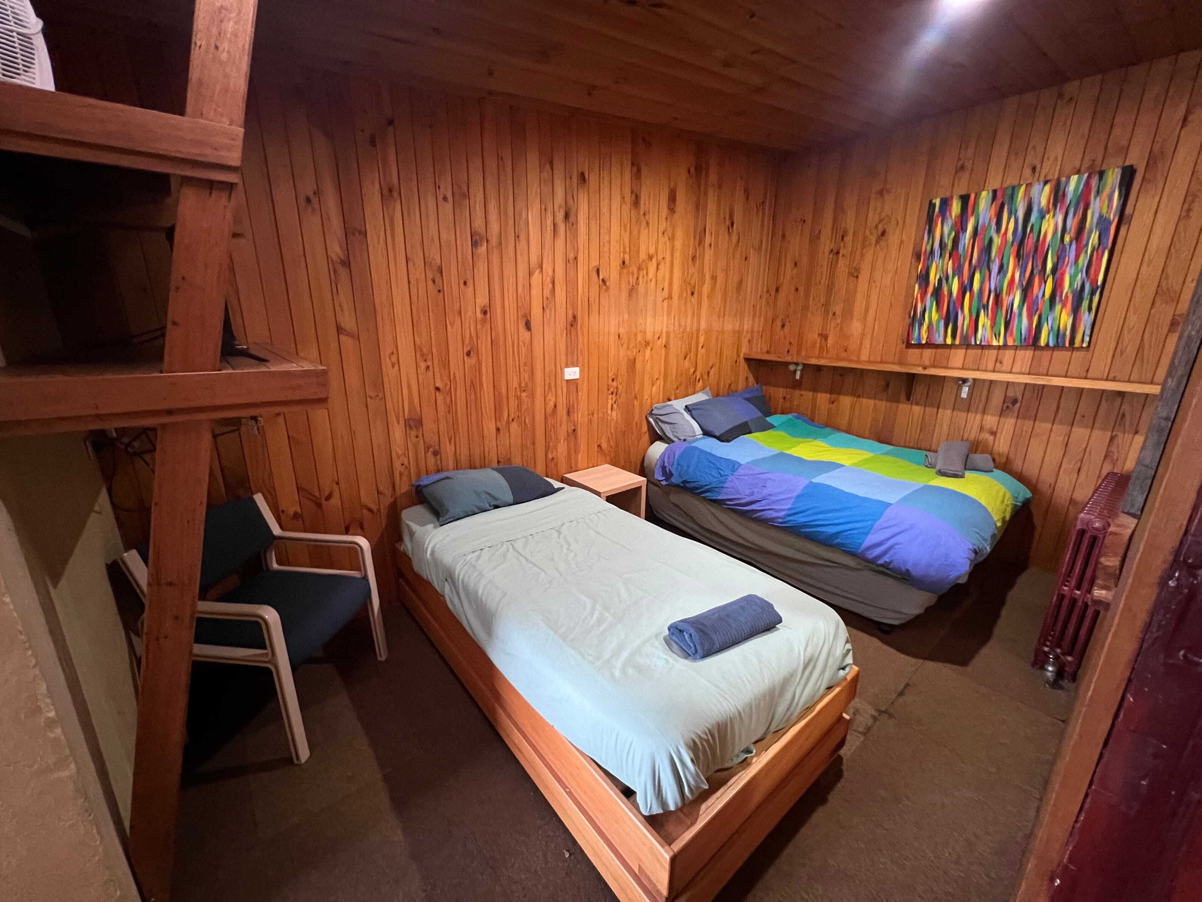 Una habitación con paneles de madera, dos camas, una silla y una obra de arte colorida en la pared.
