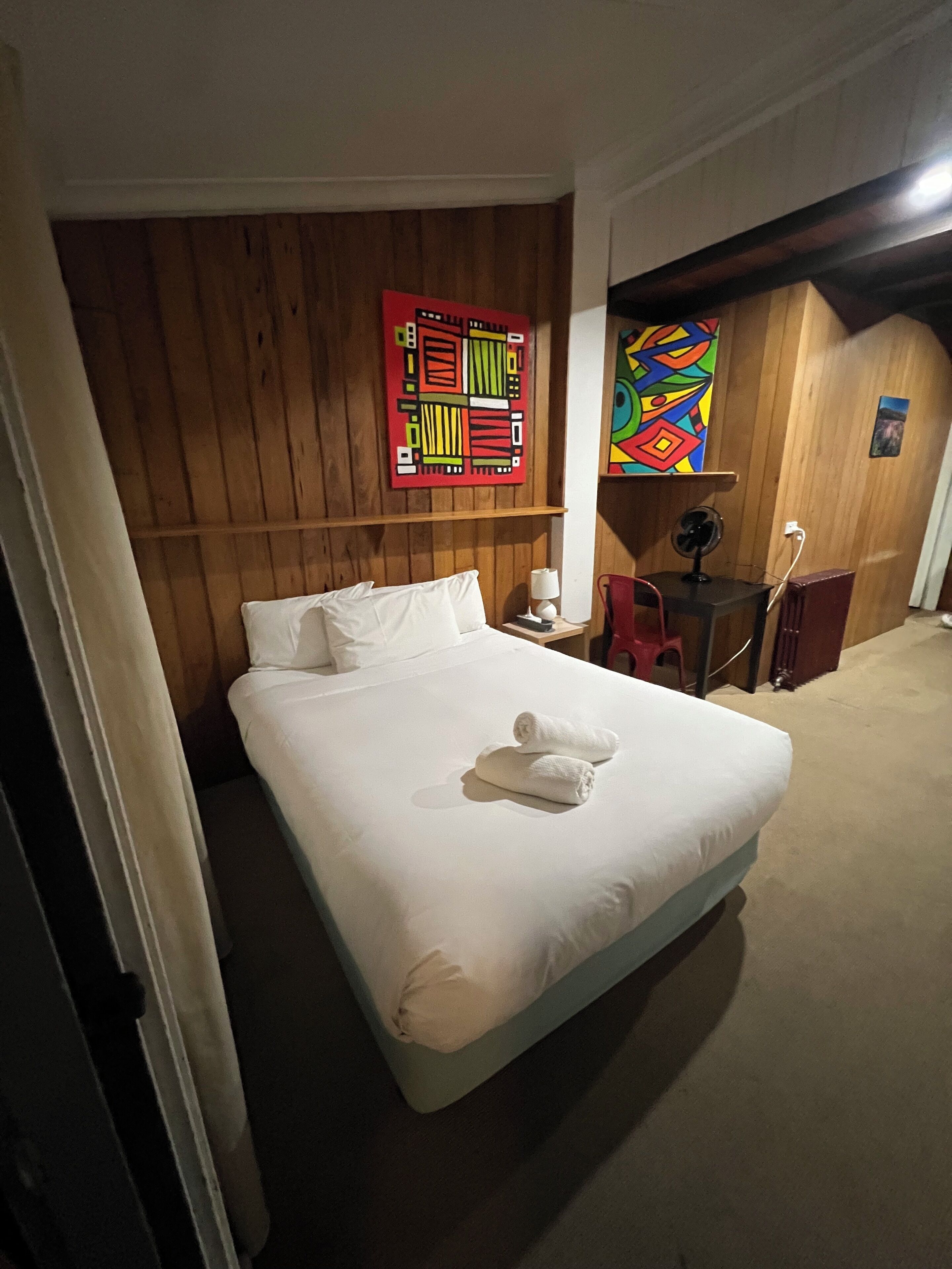 A hotel room with a bed, a desk, a chair, a fan, and two wall art pieces.