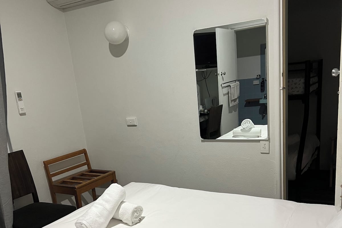 A hotel room with a bed, a chair, a small table, a mirror, and an air conditioning unit.