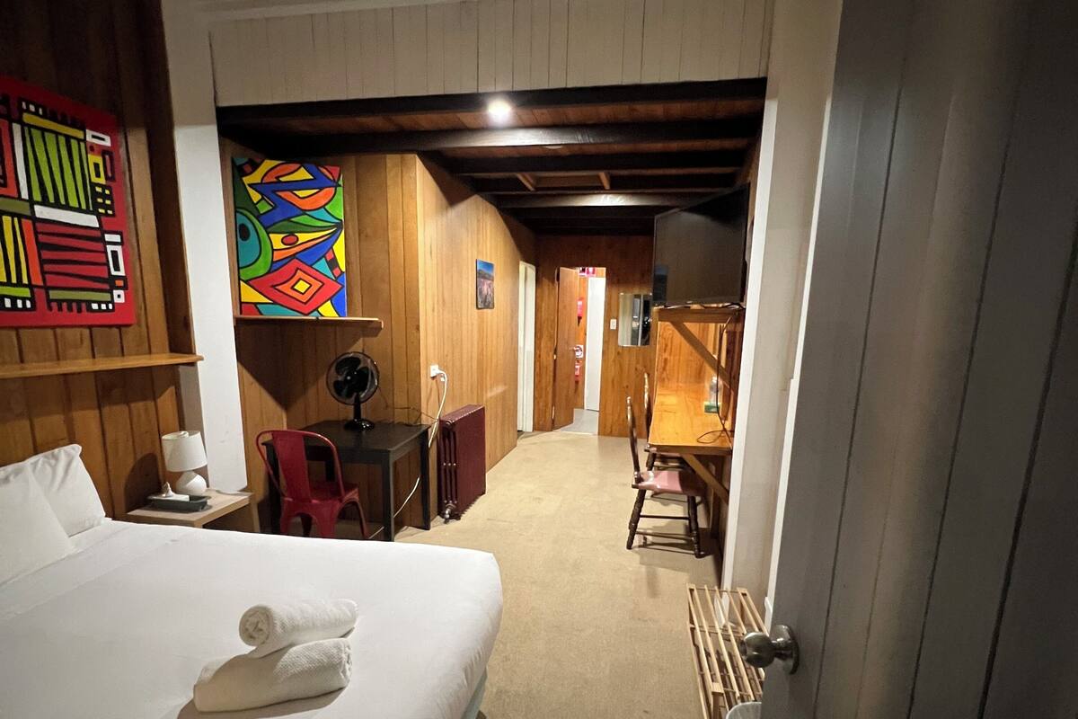 A hotel room with a bed, a desk, a chair, a lamp, and a colorful artwork on the wall.