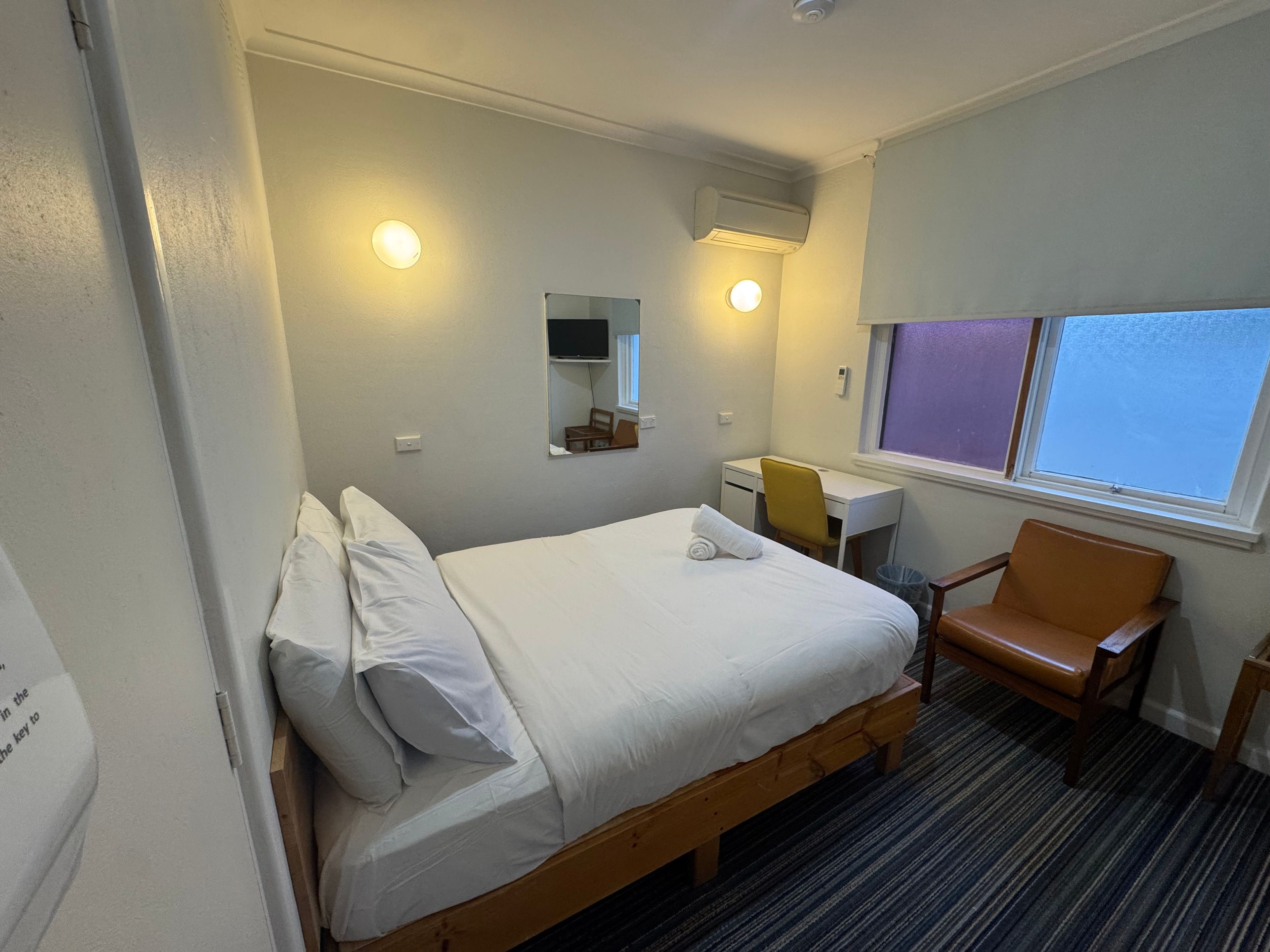 Una habitación de hotel con una cama individual, un escritorio, una silla, un ventanal y un equipo de aire acondicionado.