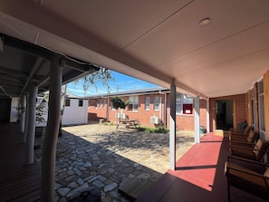A covered outdoor area with a stone floor, a wooden bench, and a brick building in the background. - Bunkhouse Motel Cooma (Cooma)