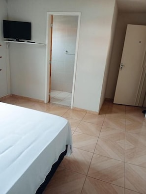 Un dormitorio con cama, televisión sobre una estantería, puerta que da a un baño y suelo de baldosas.