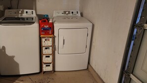 Un cuarto de lavado con dos lavadoras, una botella roja de detergente y un estante de madera con detergente.