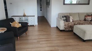 Una sala de estar con un sillón negro, un sofá color beige y un pequeño mueble blanco con un objeto decorativo encima.