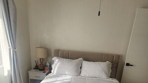 Una cama bien hecha con edredón blanco y almohadas, una mesita de noche con lámpara, un ventilador de techo y una alfombra estampada sobre piso de madera.