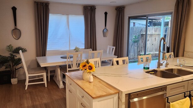 Una cocina con encimera blanca, un lavamanos y lavavajillas. Hay una mesa de comedor con sillas y una planta en maceta.