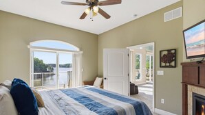 A bedroom with a bed, a ceiling fan, a fireplace, a television, and a view of the outdoors.
