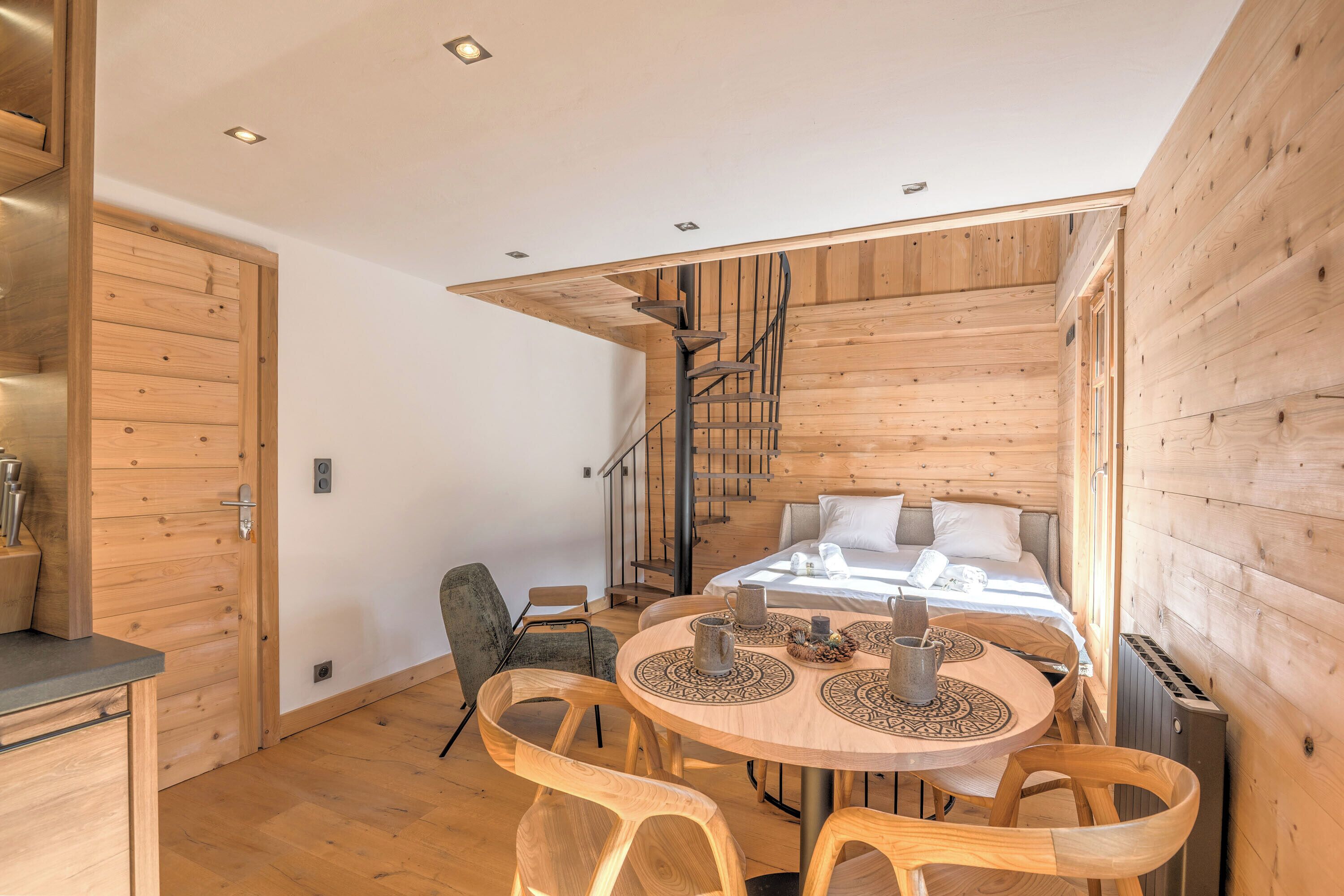 A modern log cabin with a wooden interior, a spiral staircase, a round dining table with chairs, and a bed with a white comforter.
