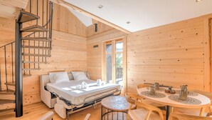 En stuga med träinredning, ett spiraltrapphus, en säng med madrass och kuddar, ett runt matbord med stolar och ett fönster som släpper in naturligt ljus.