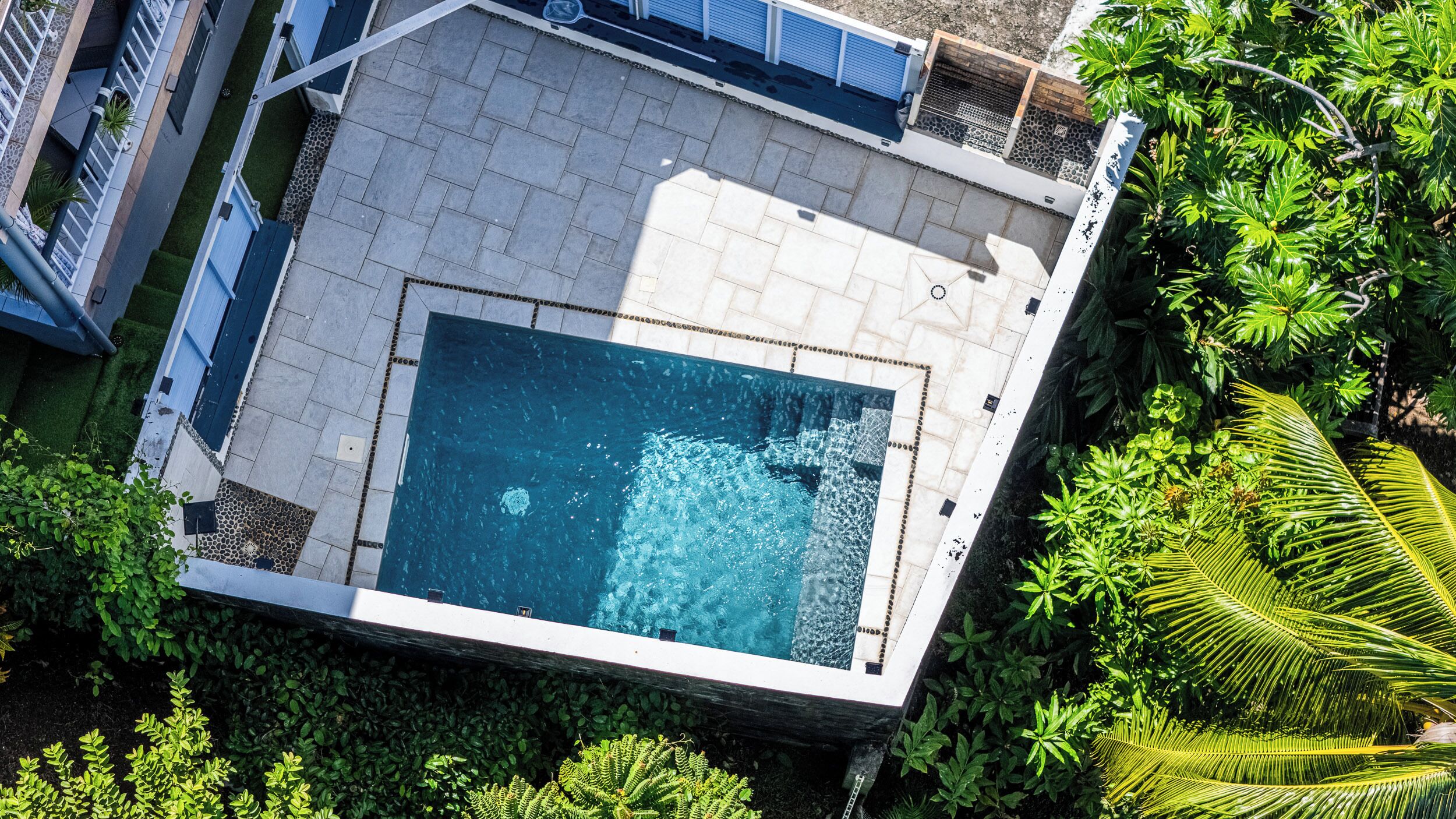 Vista aérea de una piscina moderna con diseño geométrico, rodeada de exuberante vegetación y una terraza pavimentada.