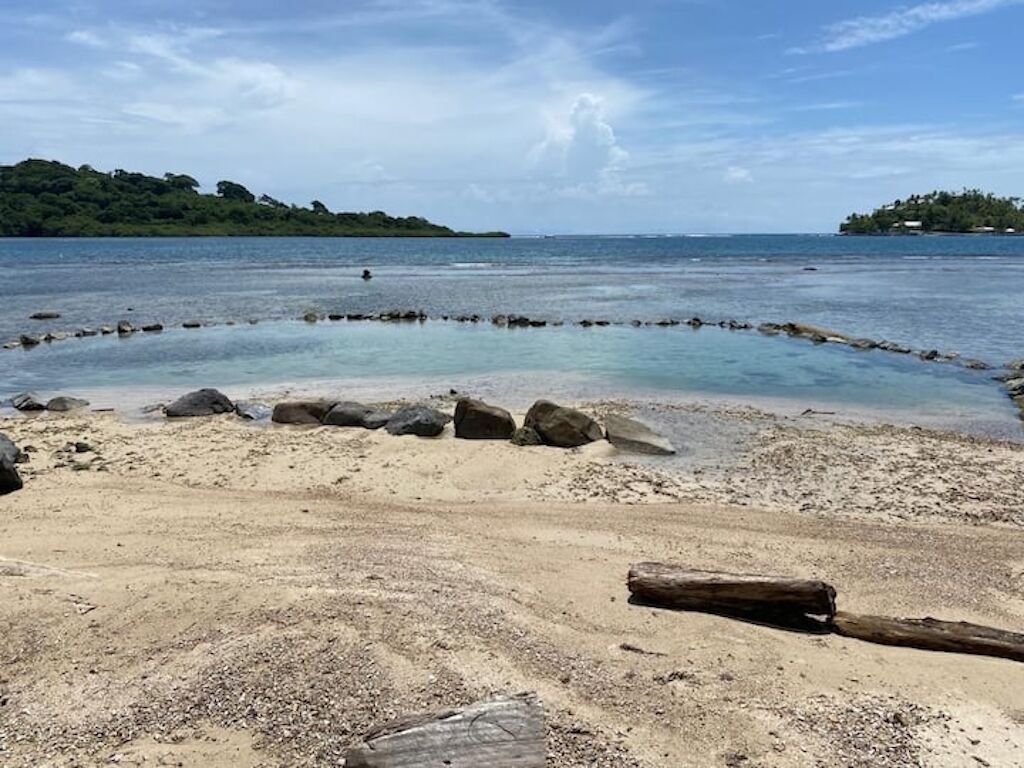 Una platja amb una formació circular de pedra, aigua tranquil·la i turons verds a la distància.