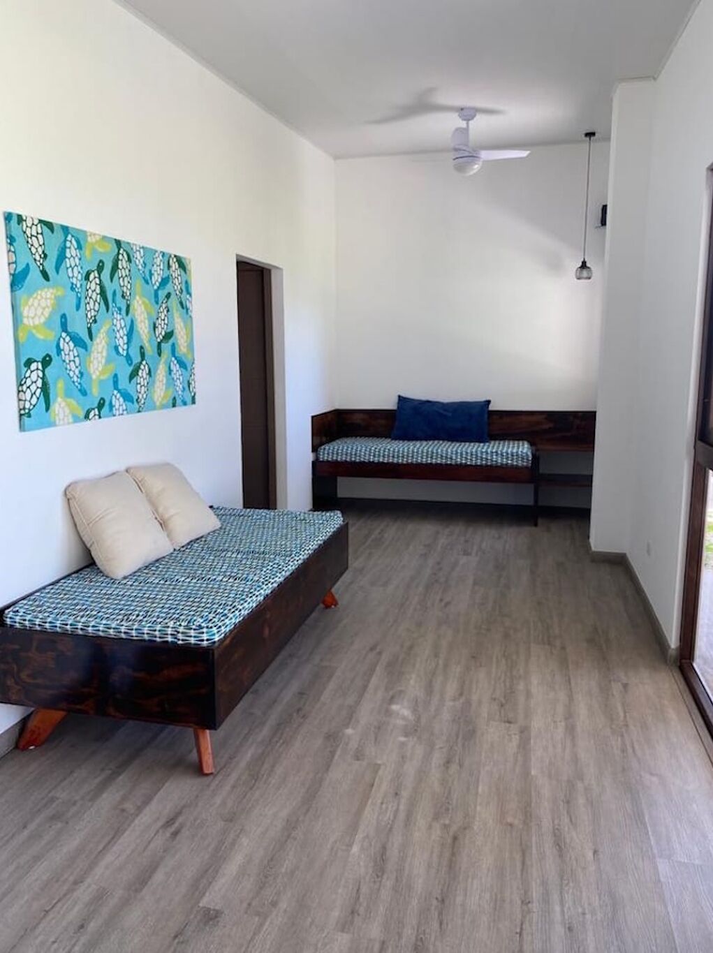 Een kamer met een houten vloer, een bed met een geruit sprei, een bankje met een blauw kussen, en een plafondventilator.
