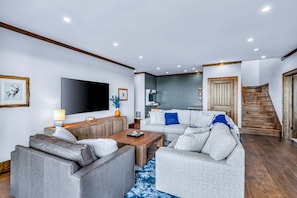 Een moderne woonkamer met een flatscreen televisie, grijze banken, een bijzettafeltje en een trap naar een houten deur.
