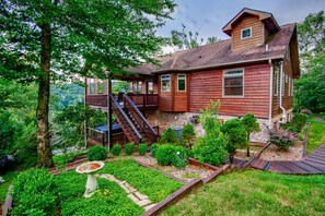 A two-story wooden house with a balcony, surrounded by a well-maintained garden with various plants and a birdbath. - Mountain Sunrise - Great Views, Hot Tub, and Game Tables on Sugar Mtn. (Banner Elk)