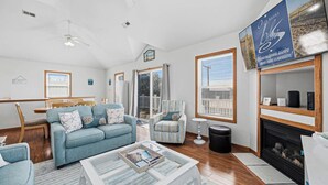 A spacious living room with a blue sofa, a coffee table, and a fireplace. There is a dining area with a wooden table and chairs. - Perfect Break | Private Pool and Hot Tub | Less than 1,000 ft to the Beach! (Kill Devil Hills)