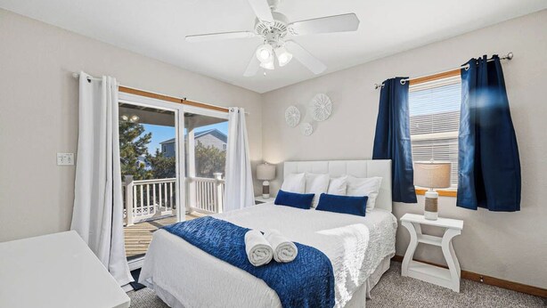 A bedroom with a bed, a nightstand, a ceiling fan, and a view of the outdoors. - Perfect Break | Private Pool and Hot Tub | Less than 1,000 ft to the Beach! (Kill Devil Hills)