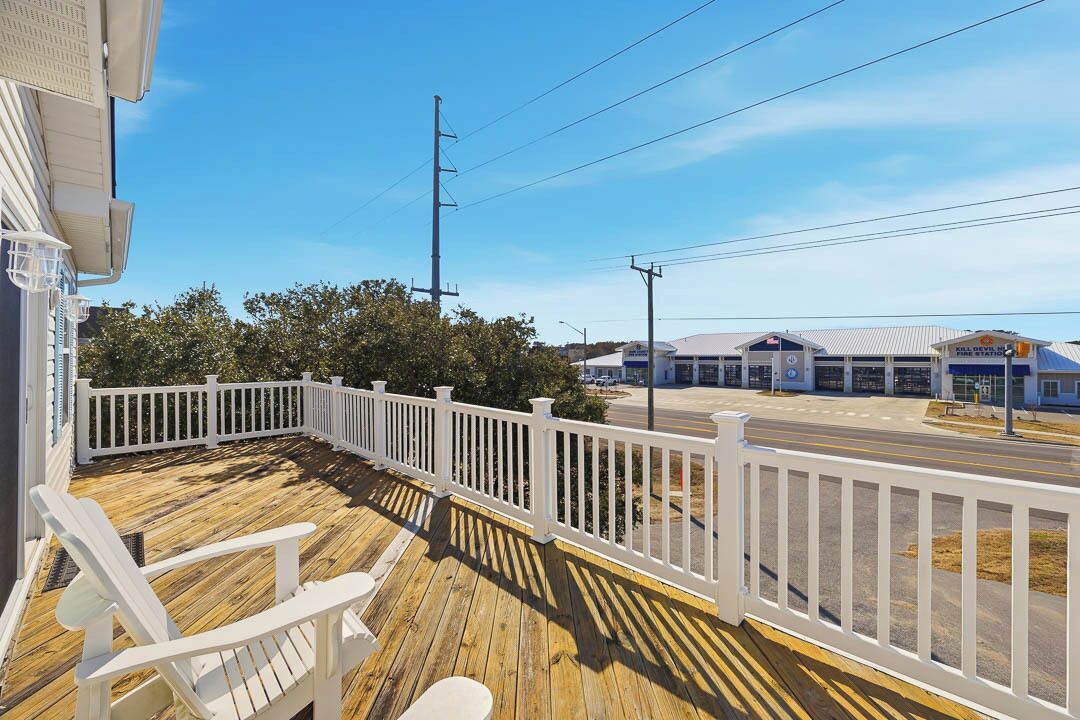 Une terrasse en bois, dotée de rampes et de chaises blanches, qui offre une vue sur une route et un bâtiment avec un toit bleu.