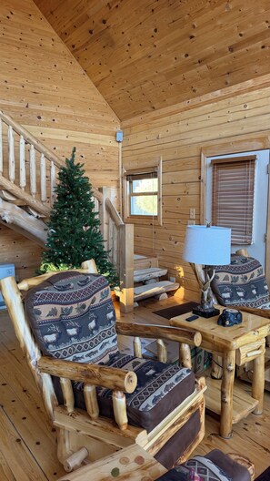 Interior de una cabaña de madera con chimenea, un árbol de Navidad, una silla de madera con cojines estampados y una mesa con una lámpara.